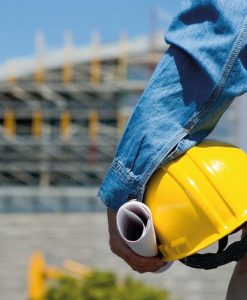Imagen en primer plano de constructor con casco amarillo en la mano, de pie frente a obra arquitectónica sin terminar, representando el técnico universitario en construcción, administración y ejecución de obra civil.