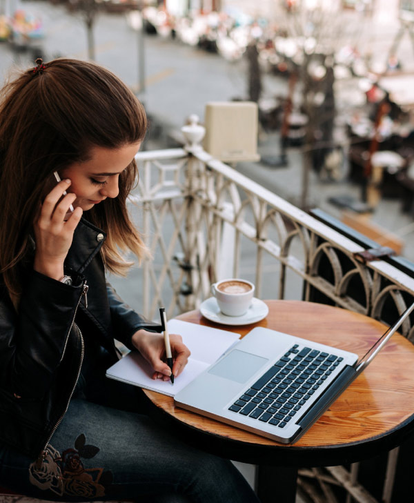 Imagen de mujer en una mesa de café, frente a una laptop escribiendo en una libreta, mientras habla por teléfono, como imagen destacada de la entrada de blog Por qué los lideres siempre están aprendiendo.